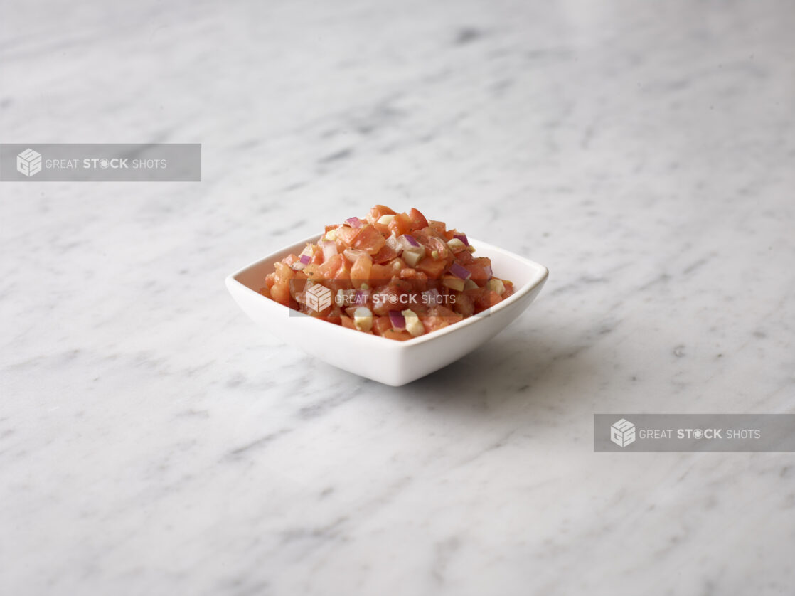 Bruschetta mix in a white square bowl on a white marble background