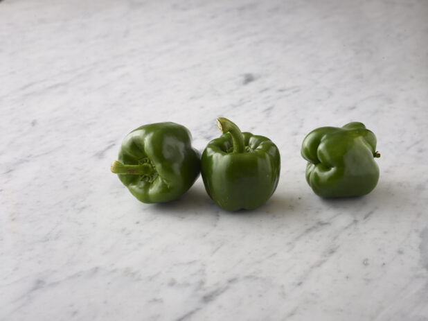 Three whole green bell peppers on a white marble background