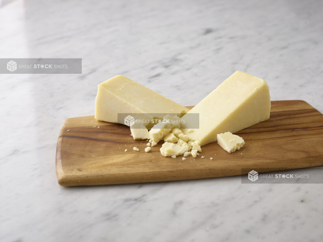 Whole wedges of cheddar cheese with crumbled pieces of cheese on a wooden board on a white marble background