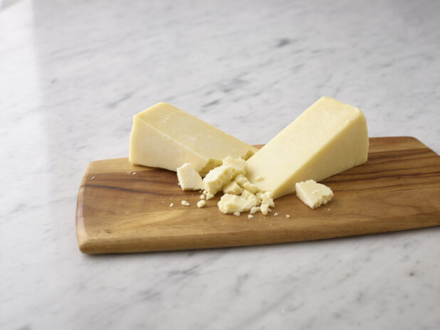 Whole wedges of cheddar cheese with crumbled pieces of cheese on a wooden board on a white marble background