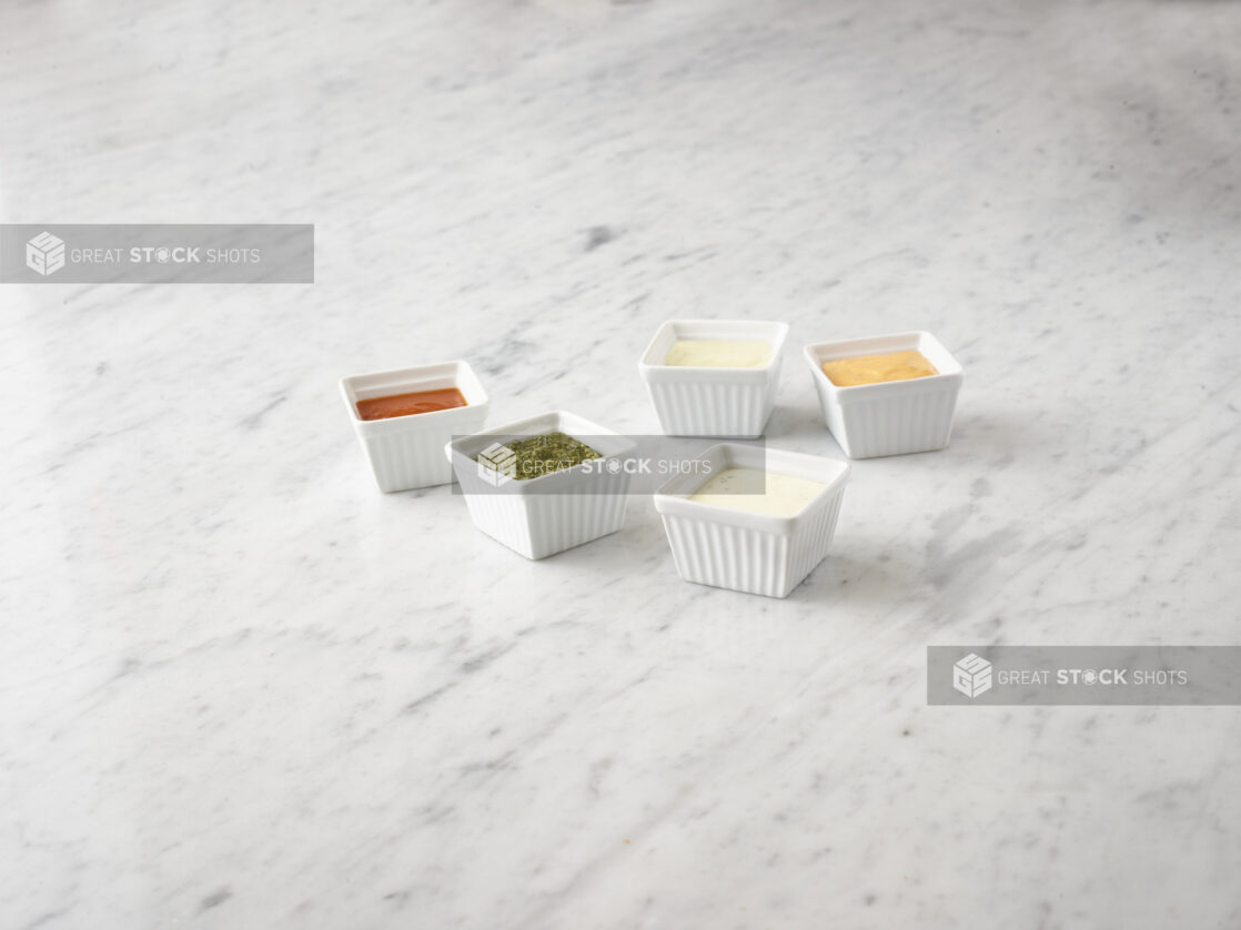 5 various condiments in white square ramekins on a white marble background