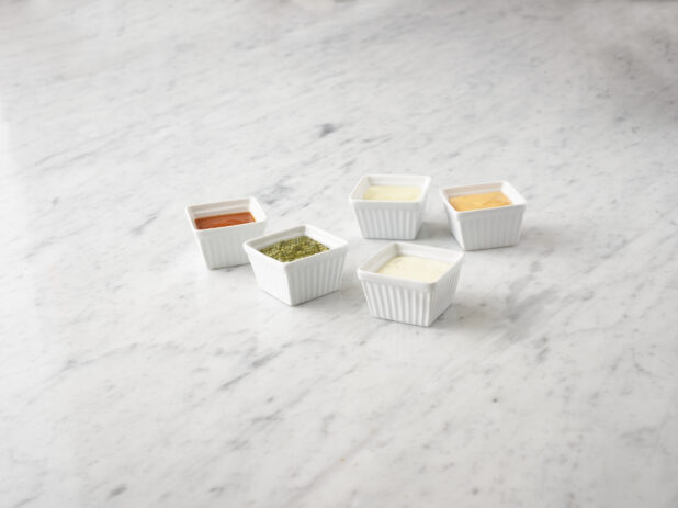 5 various condiments in white square ramekins on a white marble background