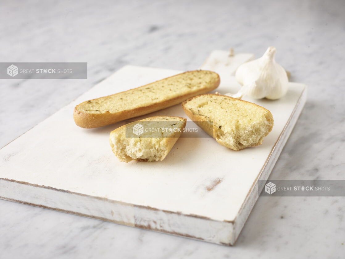 Garlic bread on a white wooden cutting board with a whole clove of garlic on a white marble background