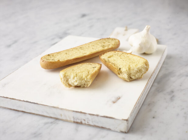 Garlic bread on a white wooden cutting board with a whole clove of garlic on a white marble background
