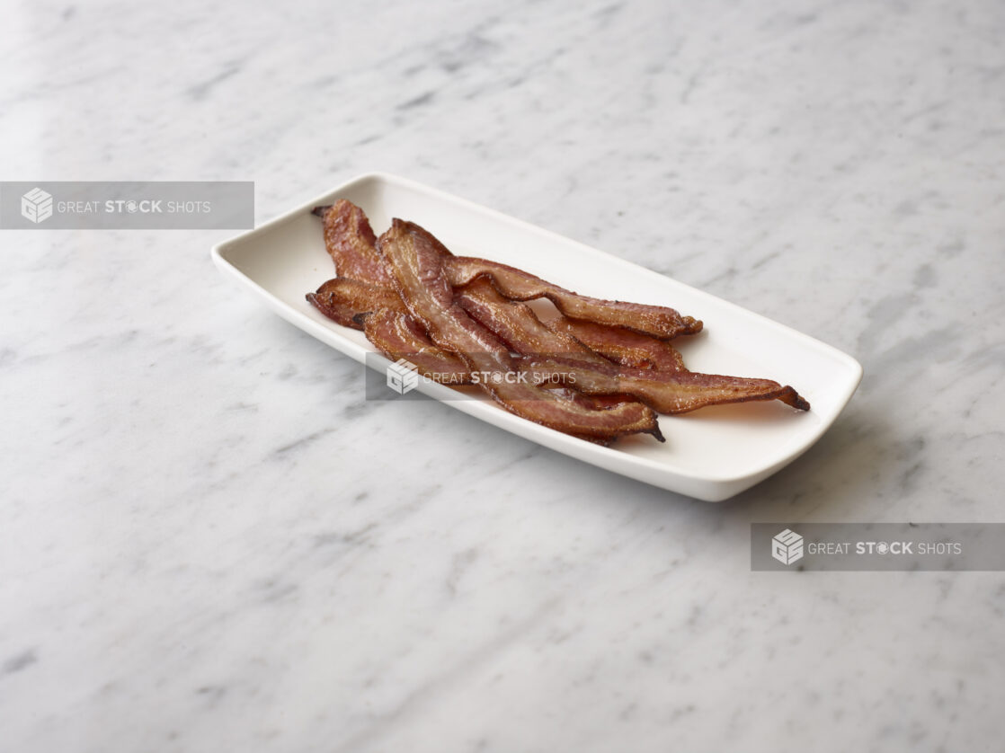 Cooked crispy sliced bacon on a rectangular white plate on a white marble background