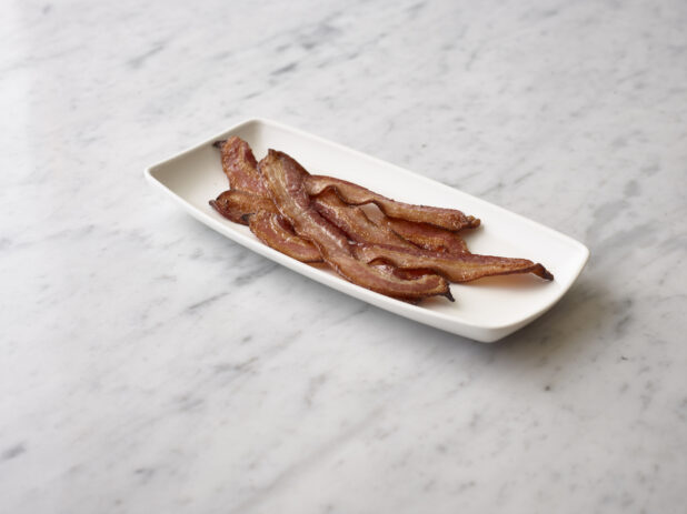 Cooked crispy sliced bacon on a rectangular white plate on a white marble background