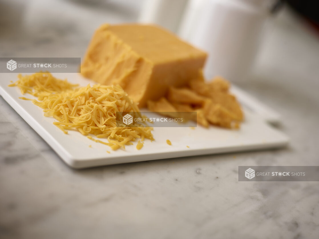 Shredded cheddar cheese with a block of cheddar and crumbled cheddar on a white cutting board on a white marble background