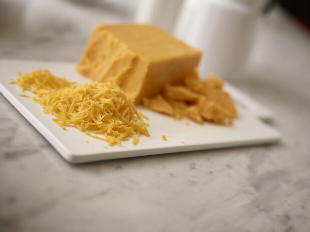 Shredded cheddar cheese with a block of cheddar and crumbled cheddar on a white cutting board on a white marble background