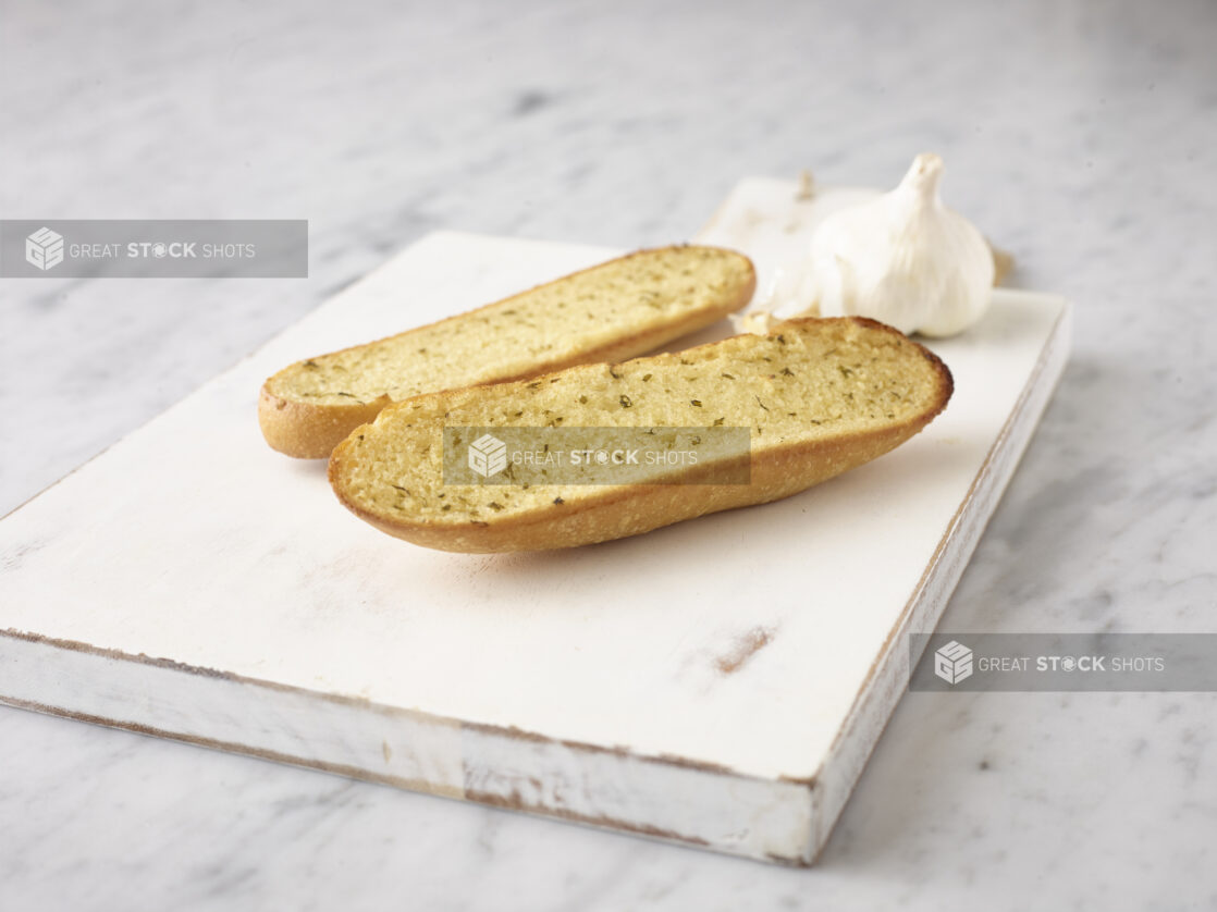 2 slices of garlic bread on a white board with whole garlic bulb in the background, on a white marble background