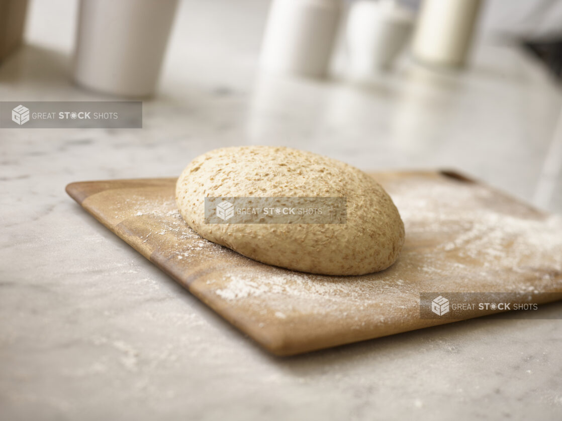 Raw whole wheat pizza dough on a wooden board with flour underneath and around the board, on a white marble background