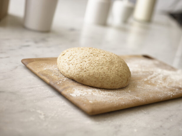Raw whole wheat pizza dough on a wooden board with flour underneath and around the board, on a white marble background