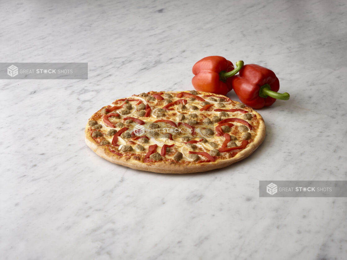 Whole 3 topping pizza with sausage, onions and roasted red peppers with 2 whole fresh red bell peppers in the background on a white marble background