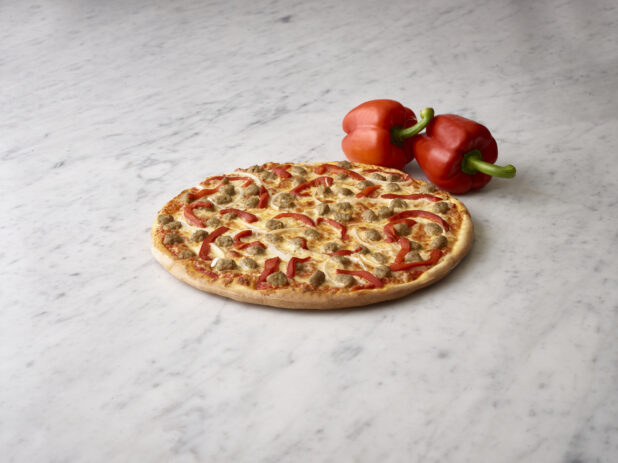 Whole 3 topping pizza with sausage, onions and roasted red peppers with 2 whole fresh red bell peppers in the background on a white marble background