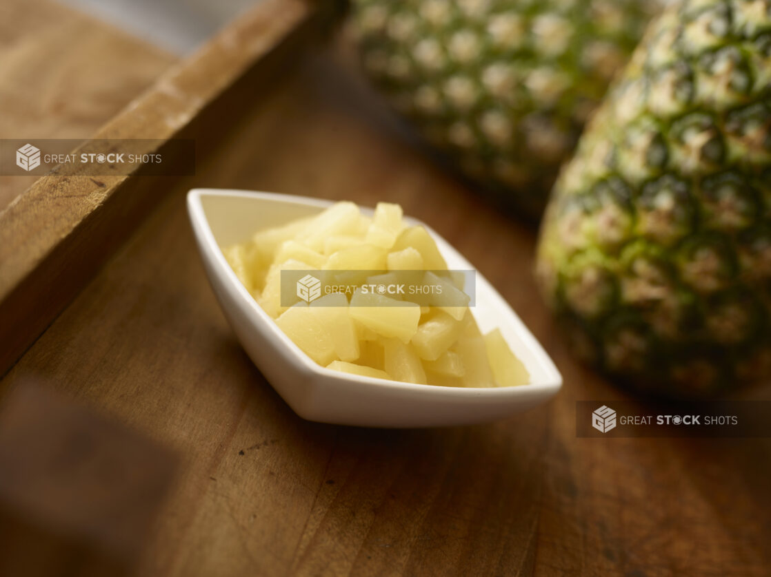 Pineapple pieces in a white side square bowl in the foreground with whole pineapple in the background all on a rustic wooden background