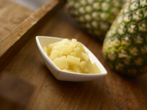 Pineapple pieces in a white side square bowl in the foreground with whole pineapple in the background all on a rustic wooden background