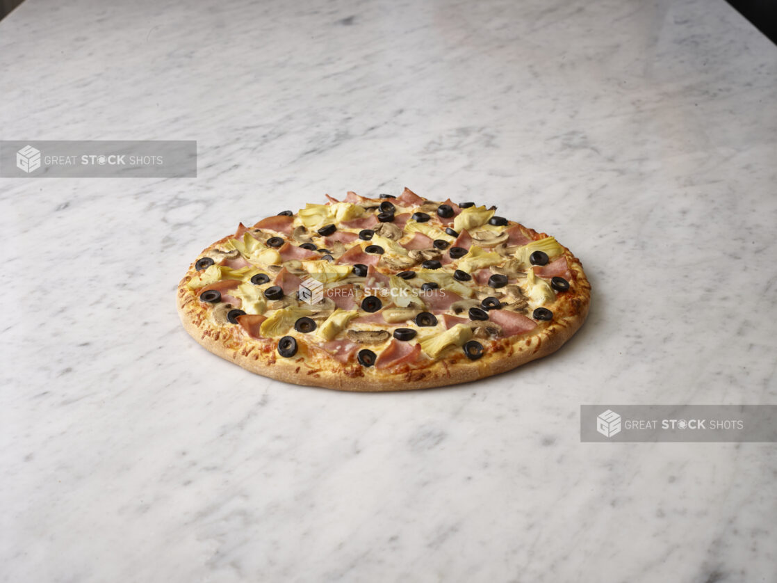 Whole unsliced artichoke, black olive, and ham pizza on a white marble background