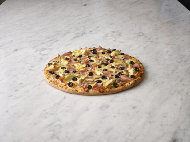 Whole unsliced artichoke, black olive, and ham pizza on a white marble background