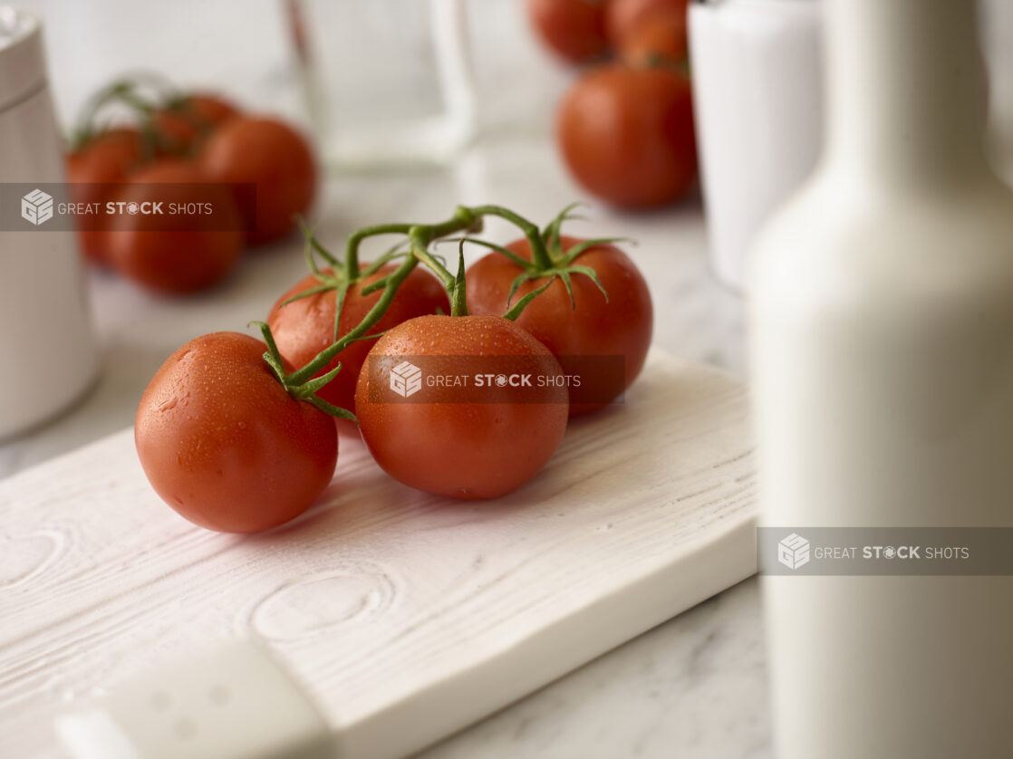 Whole tomatoes on the vine on a white board in the foreground with whole tomatoes in the background all on a white marble background