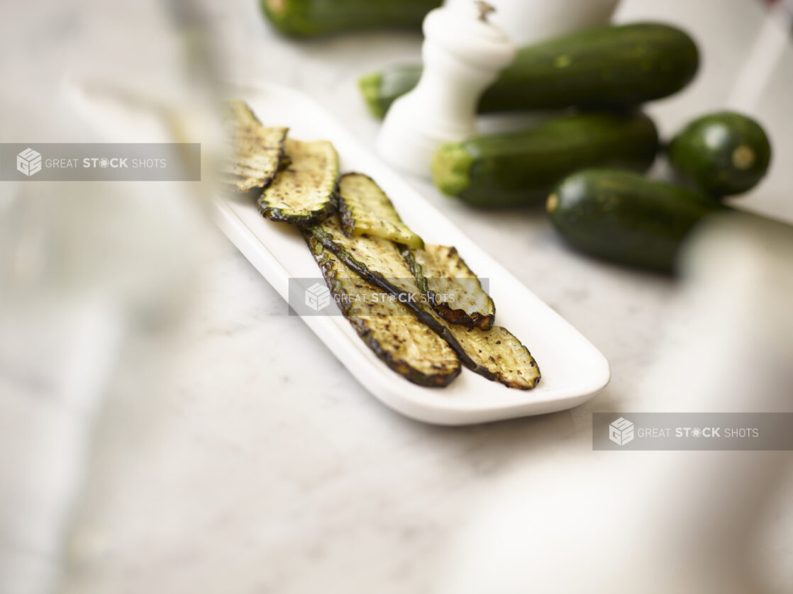 Grilled green zucchini on a white oval platter with whole green zucchini in the background on a white marble background