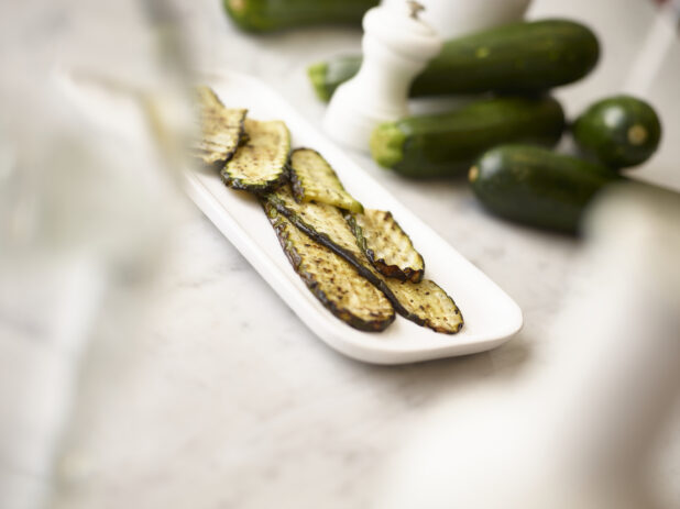 Grilled green zucchini on a white oval platter with whole green zucchini in the background on a white marble background