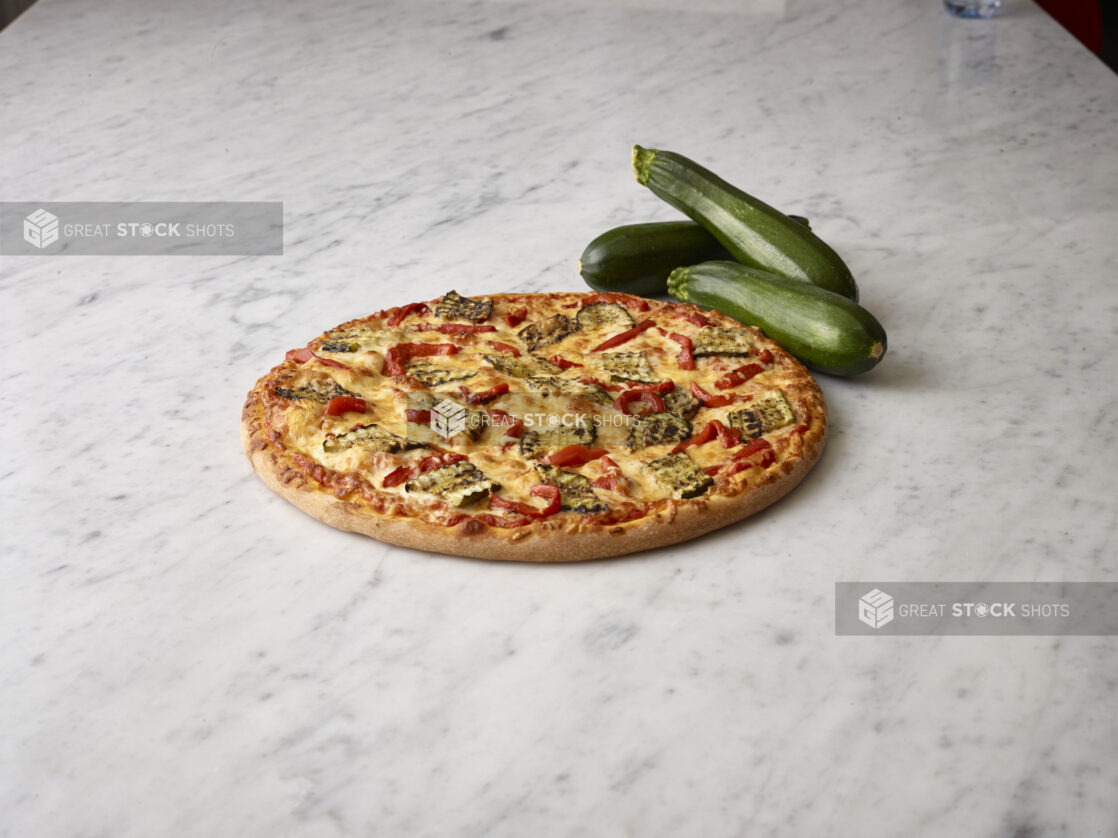 Whole vegetarian pizza with grilled zucchini and roasted red peppers with whole green zucchini in the background all on a white marble background