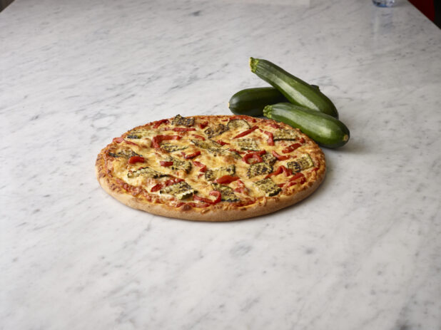 Whole vegetarian pizza with grilled zucchini and roasted red peppers with whole green zucchini in the background all on a white marble background