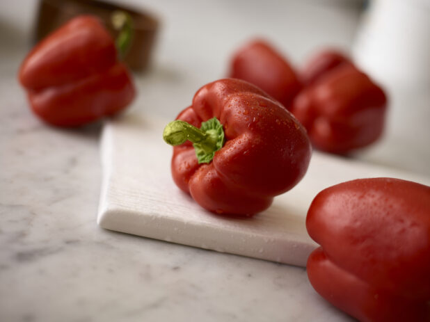 Whole red bell peppers in the foreground and the background, on a white board on a white marble background