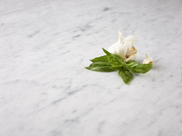 Off center view of fresh basil and a fresh garlic bulb with cloves on a white marble background