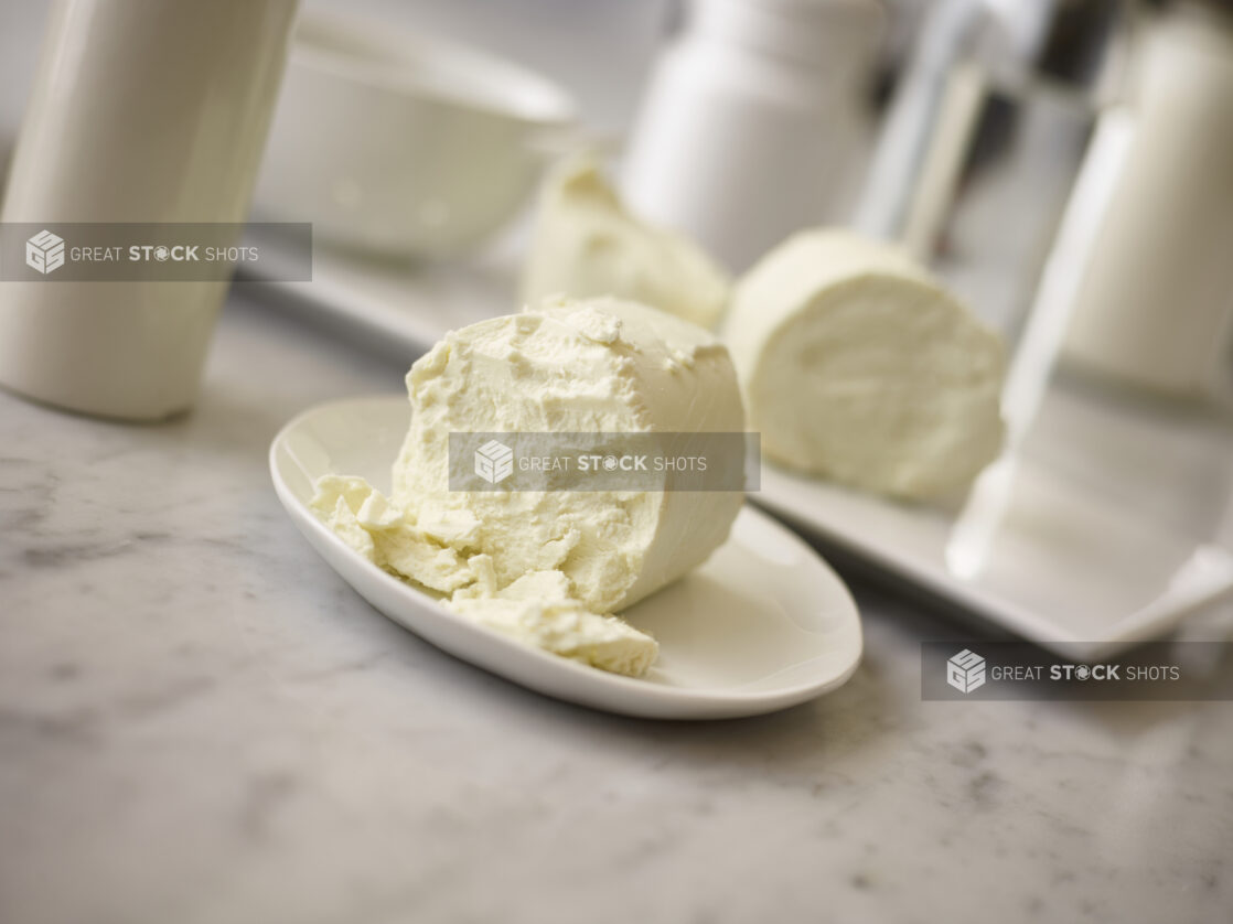 Goat cheese on a small round white plate in the foreground with goat cheese and accessories in the background on a white marble background