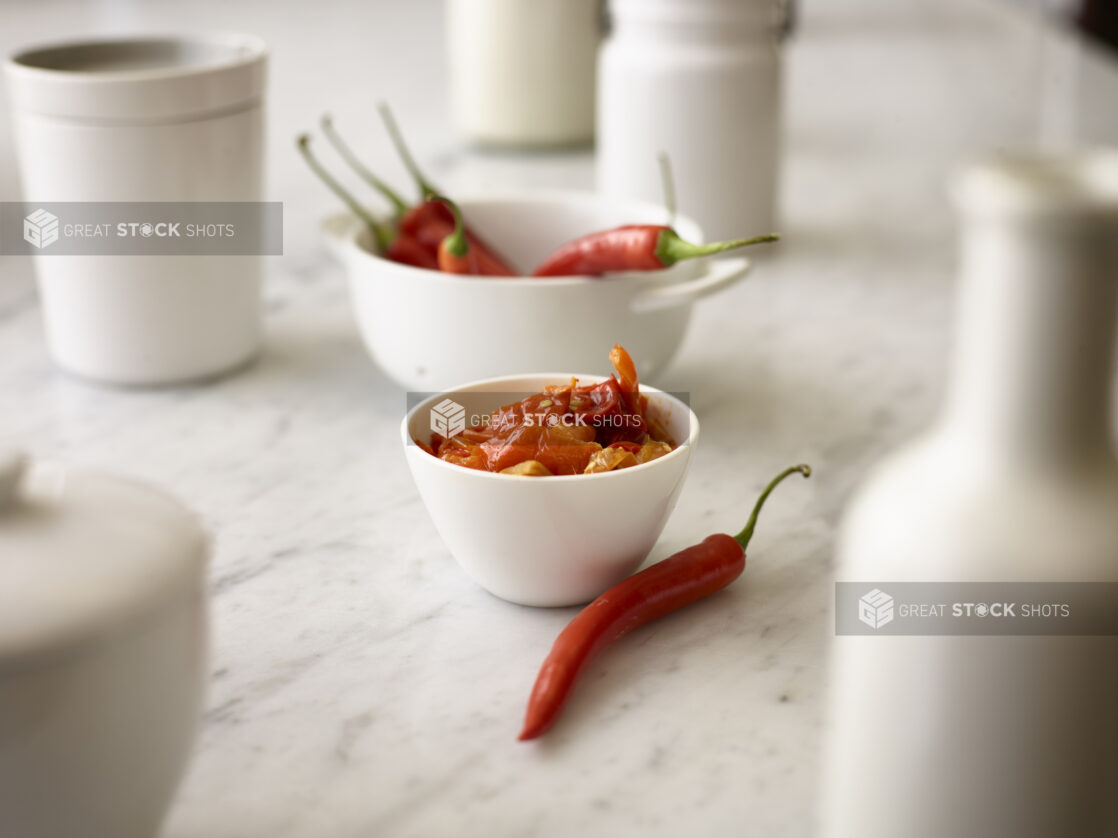 Roasted red chilis in a small round white bowl with a whole fresh red chili in the foreground and whole fresh red chilis in a white round bowl in the background all on a white marble background