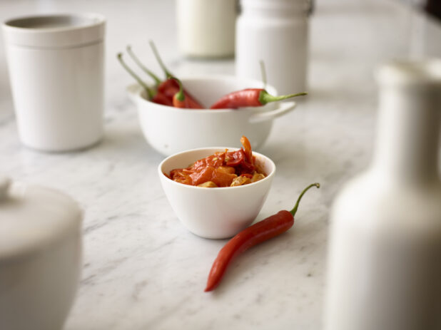 Roasted red chilis in a small round white bowl with a whole fresh red chili in the foreground and whole fresh red chilis in a white round bowl in the background all on a white marble background