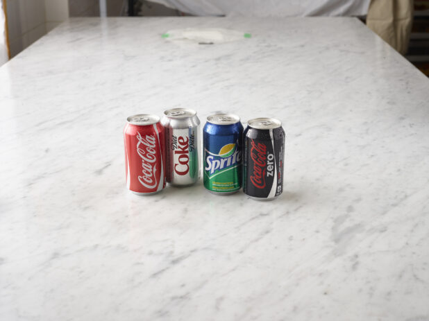 1 can of Coke, 1 can of Diet Coke, 1 can of Sprite and 1 Can of Coke Zero all on a white marble background