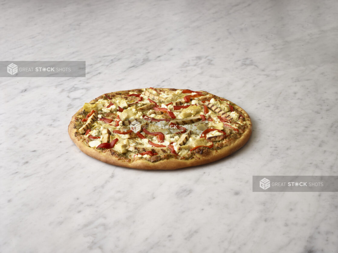 Whole pizza with chicken, artichokes, roasted red peppers and feta cheese on a white marble background