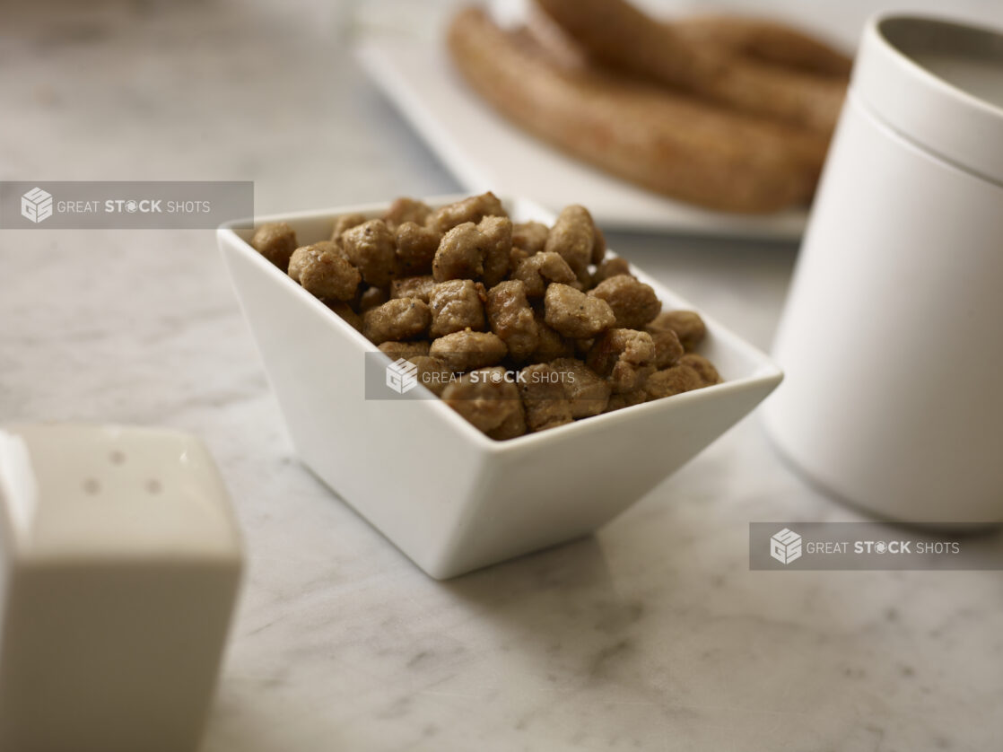 Cooked Italian sausage, pizza topping, in a white square bowl on a 45 degree angle on a white marble background