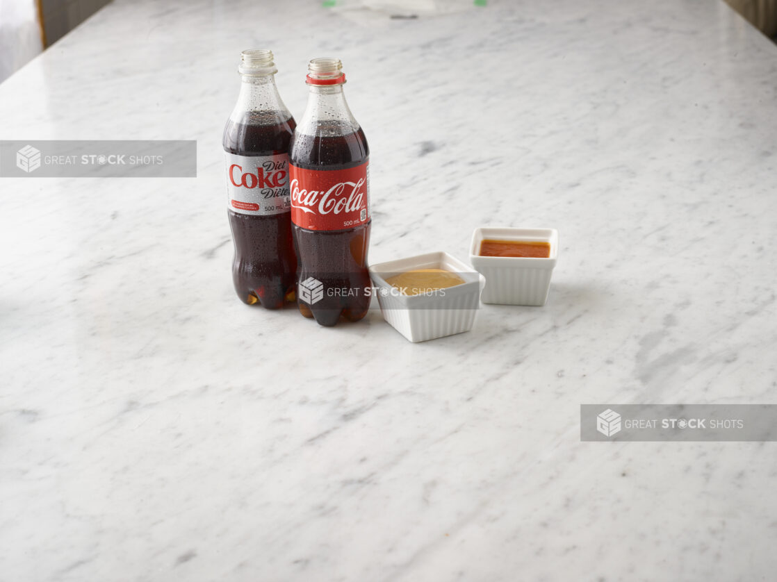 1 bottle of Coke and 1 bottle of Diet Coke with 2 white ramekins of dipping sauces on a white marble background