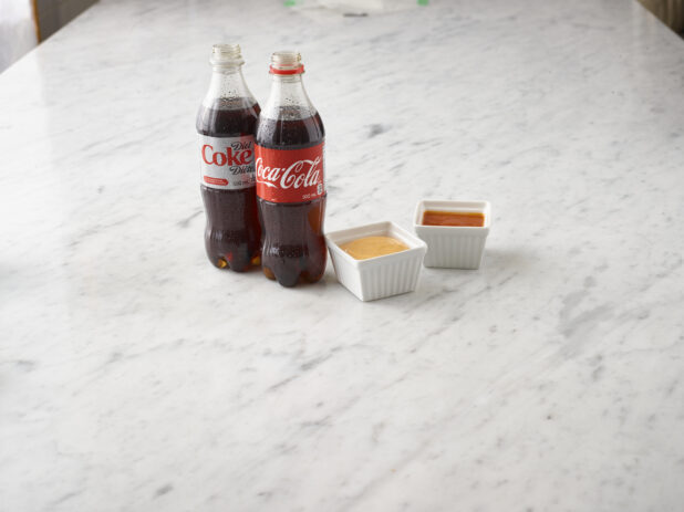 1 bottle of Coke and 1 bottle of Diet Coke with 2 white ramekins of dipping sauces on a white marble background