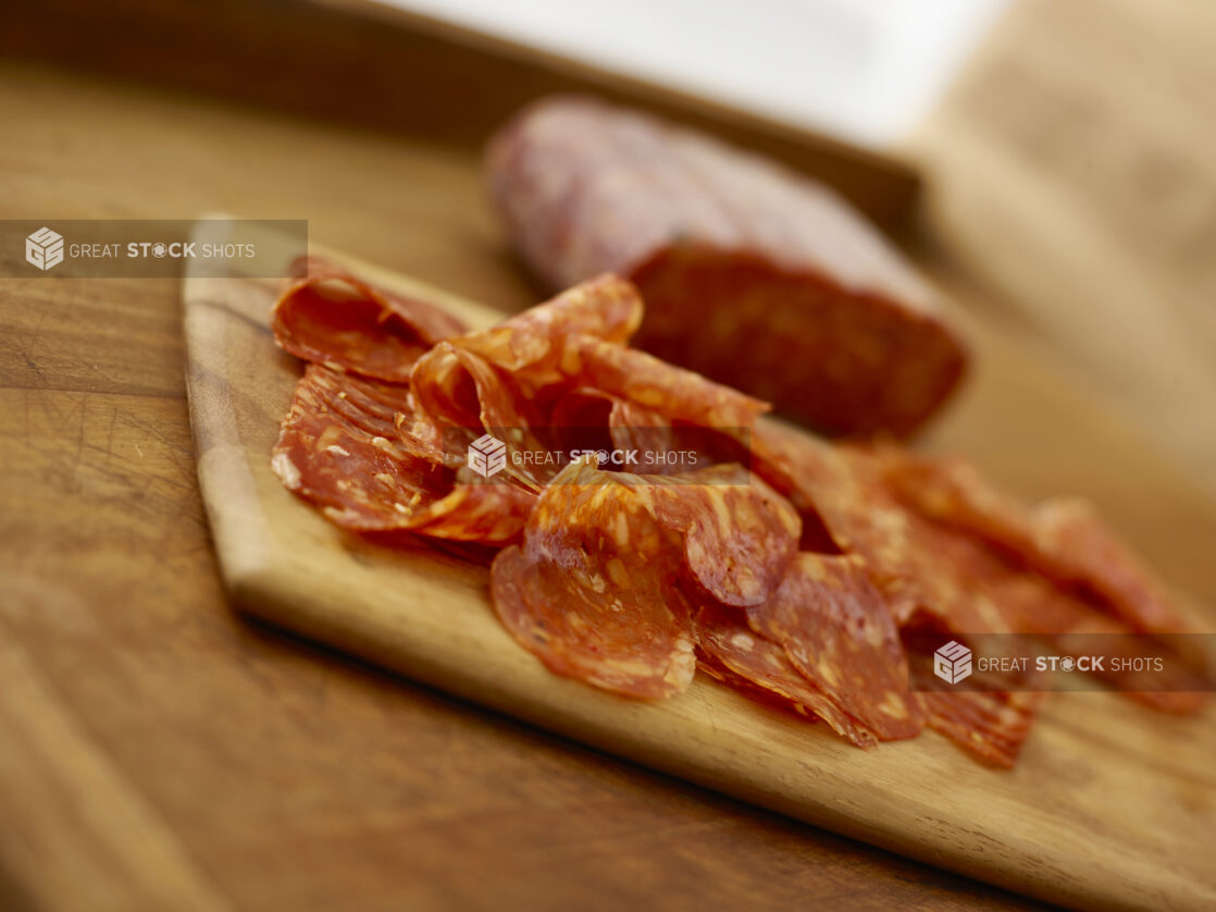 Sliced salami on a wooden cutting board in the foreground and a whole piece of salami on the background all on a rustic wooden table