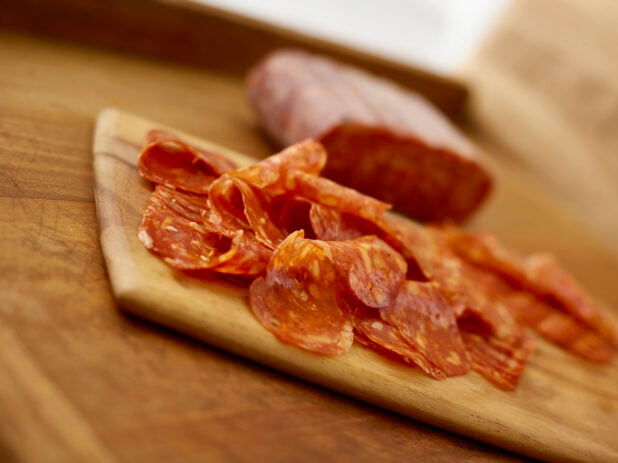 Sliced salami on a wooden cutting board in the foreground and a whole piece of salami on the background all on a rustic wooden table