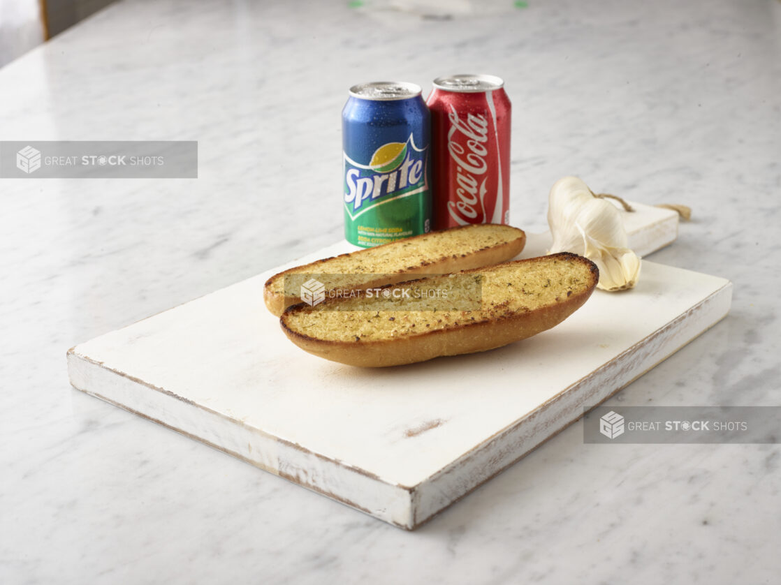 2 pieces garlic bread on a white wooden cutting board with a can of Coke and Sprite and whole garlic clove in the background all on a white marble background