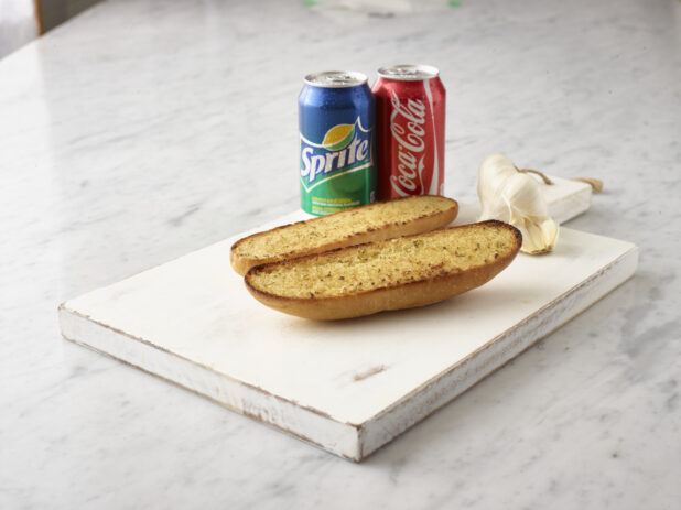 2 pieces garlic bread on a white wooden cutting board with a can of Coke and Sprite and whole garlic clove in the background all on a white marble background