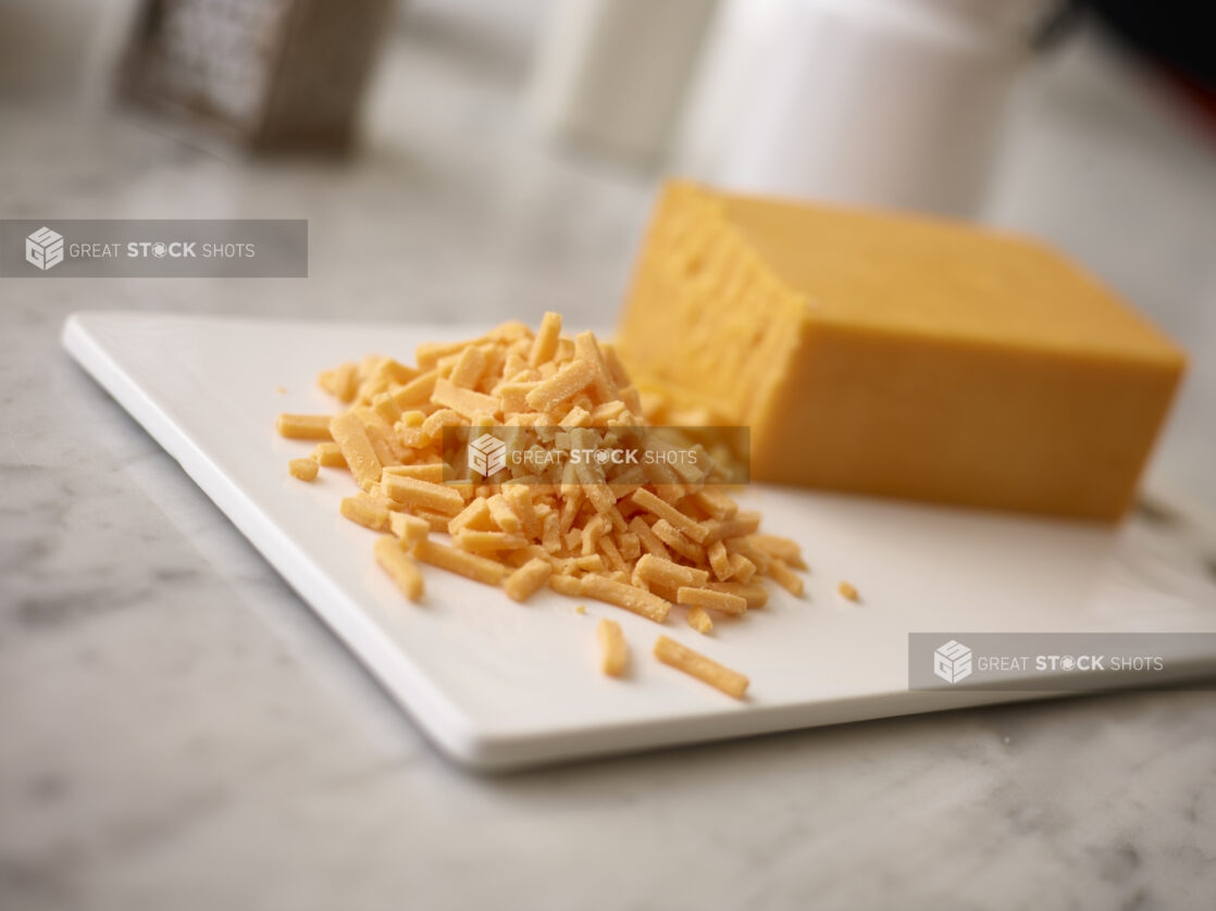 Brick of cheddar cheese on a white cutting board in the background with shredded cheddar in the foreground on a white marble background