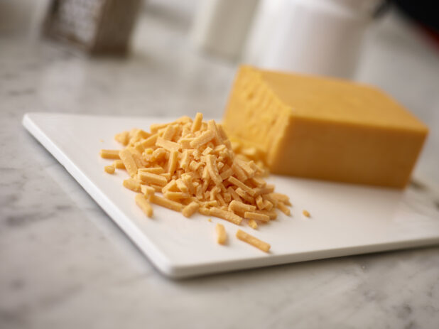 Brick of cheddar cheese on a white cutting board in the background with shredded cheddar in the foreground on a white marble background