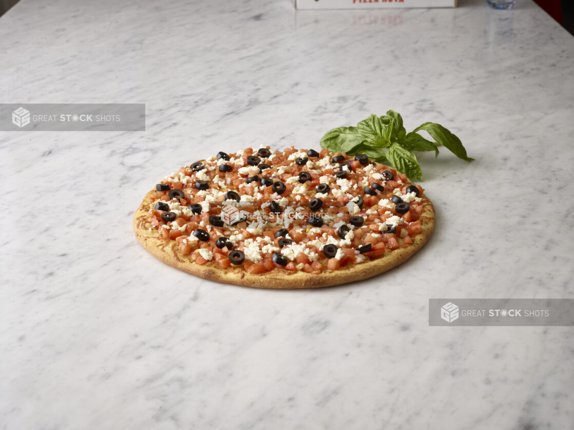 Whole unsliced chopped tomato, black olive, and feta pizza with sprig of basil alongside on a white marble background