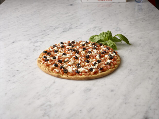 Whole unsliced chopped tomato, black olive, and feta pizza with sprig of basil alongside on a white marble background