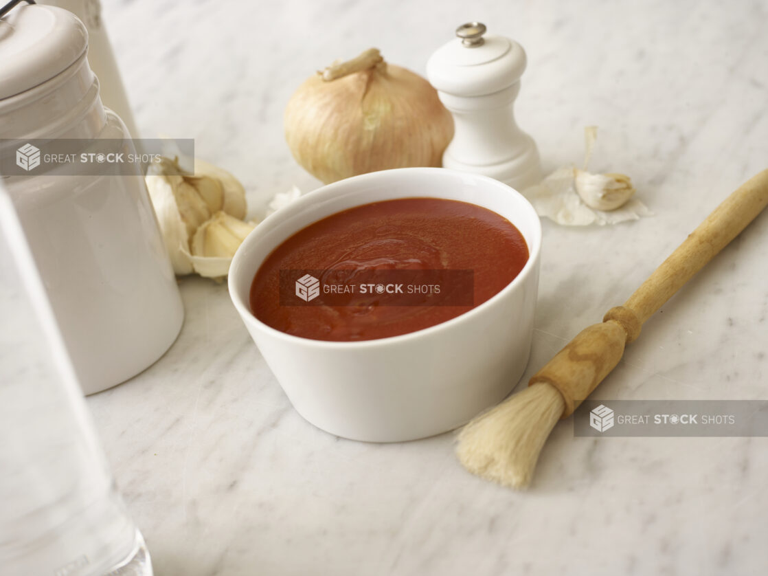 Tomato sauce in a small white bowl surrounded by a basting brush, whole garlic cloves, onion and white accessories on a white marble table