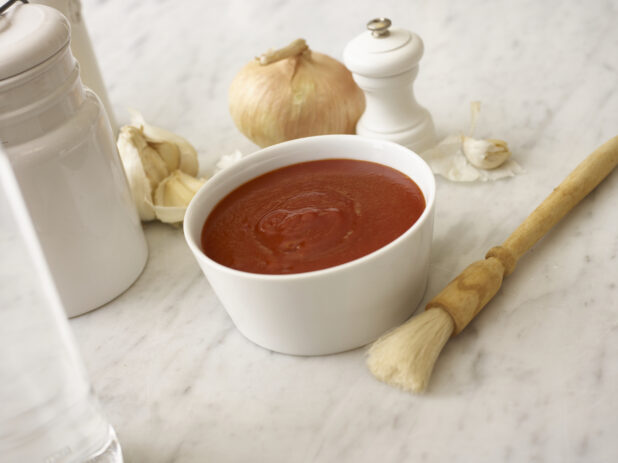Tomato sauce in a small white bowl surrounded by a basting brush, whole garlic cloves, onion and white accessories on a white marble table