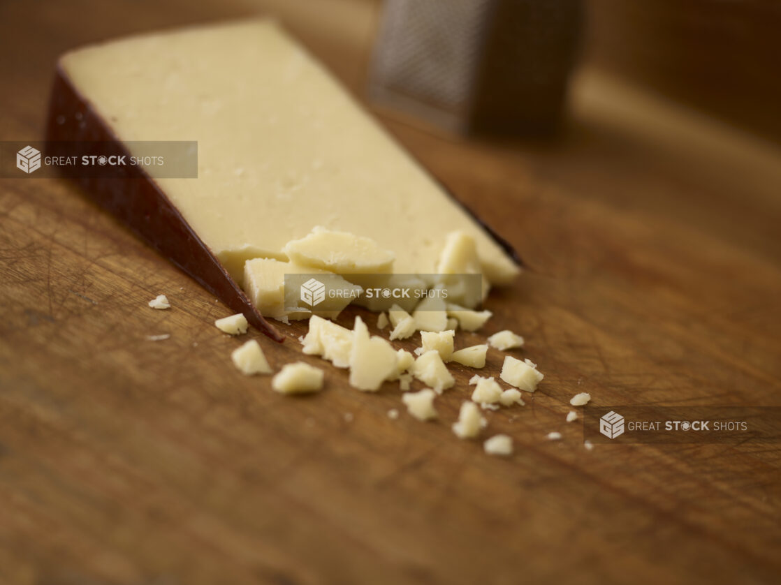 Wedge of cheese with some crumble cheese in the foreground and a cheese grater in the background all on a wooden background