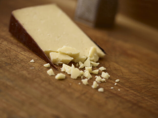 Wedge of cheese with some crumble cheese in the foreground and a cheese grater in the background all on a wooden background