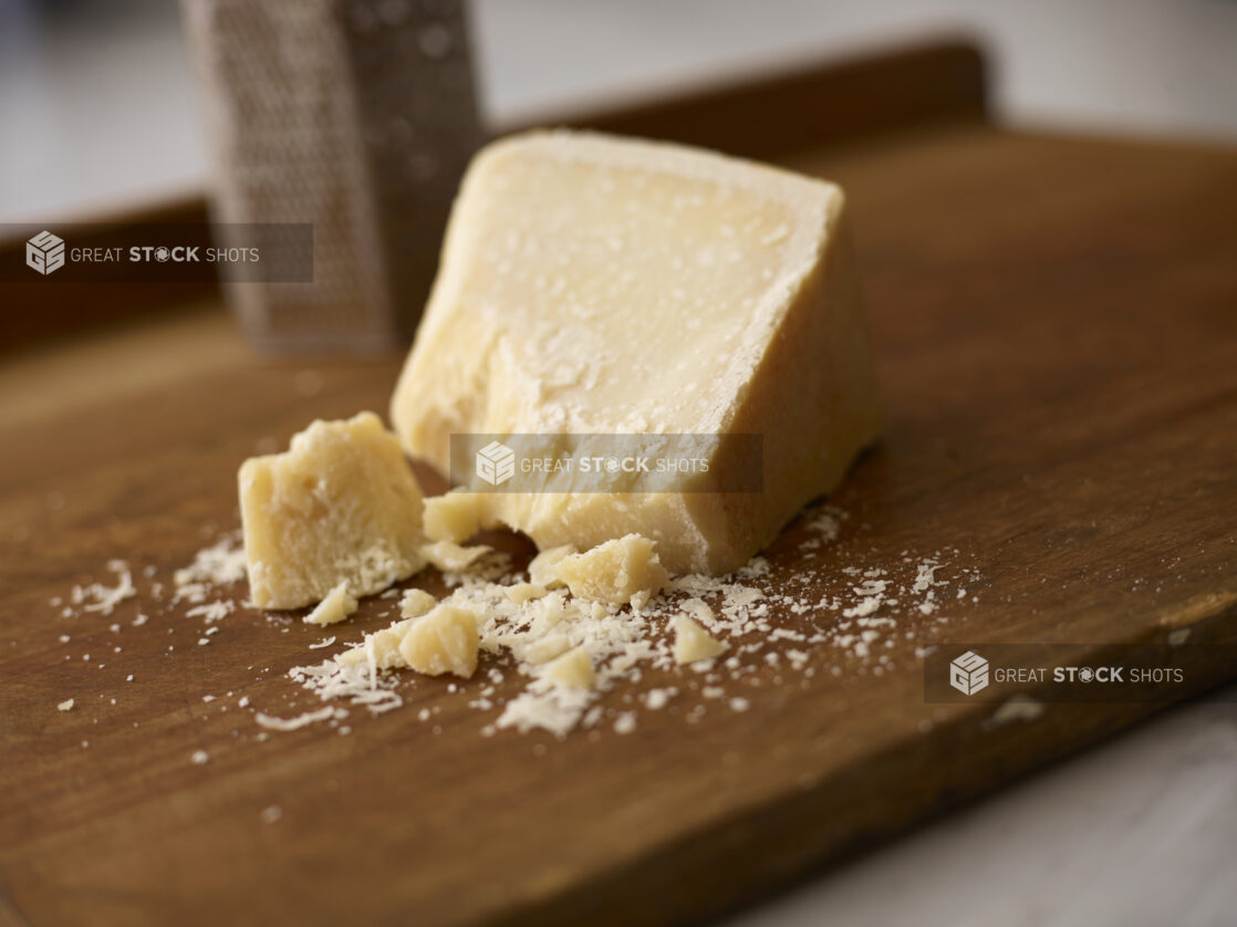 Whole parmesan cheese on a wooden board with pieces of parmesan in the forefront with a cheese grater in the background