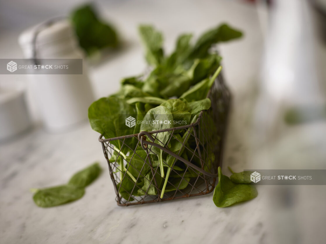 Whole fresh spinach in a decorative wire basket on a white marble table with white accessories in the background